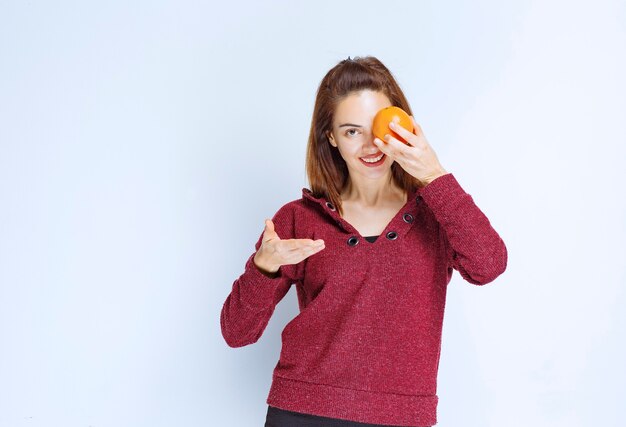 Девушка в красной куртке держит апельсин перед глазом.
