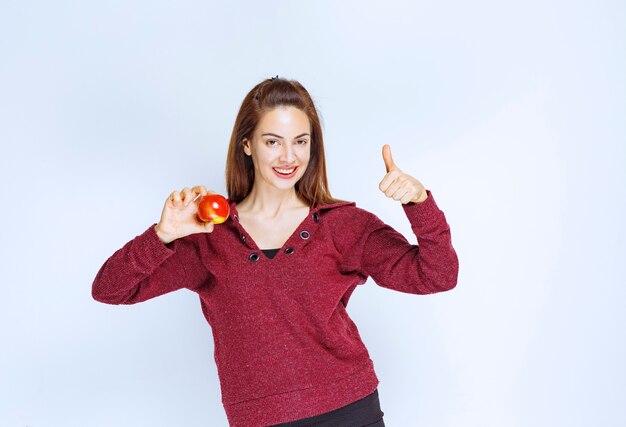 Girl in red jacket holding a red apple and showing positive hand sign