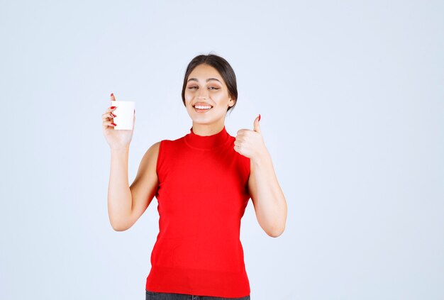 Девушка в красной рубашке пьет кофе и показывает положительный знак.