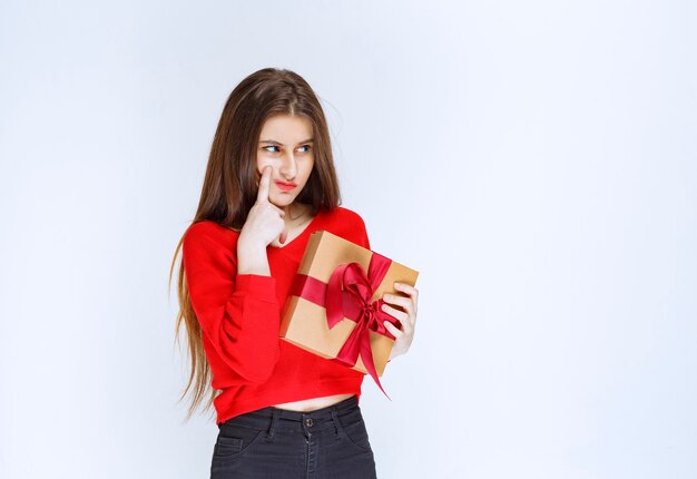 Girl in red shirt holding a cardboard gift box wrapped with red ribbon and looks confused and thoughtful. 