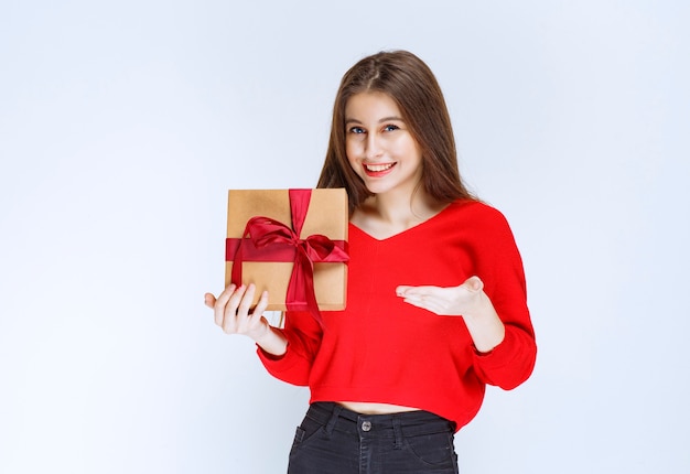 Ragazza in camicia rossa che tiene una scatola regalo di cartone avvolta con nastro rosso.