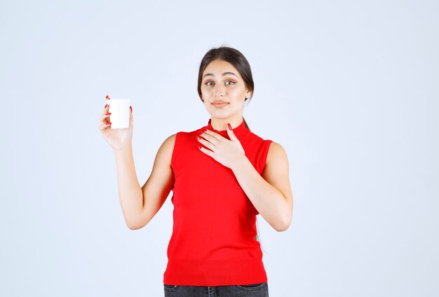 Girl in red shirt holding a white coffee cup and pointing at herself.