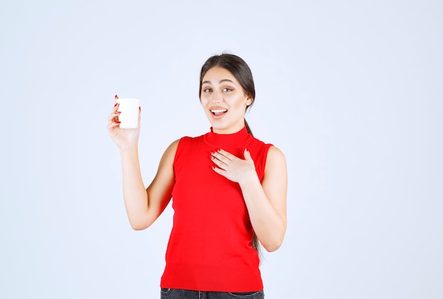 Girl in red shirt holding a white coffee cup and pointing at herself.