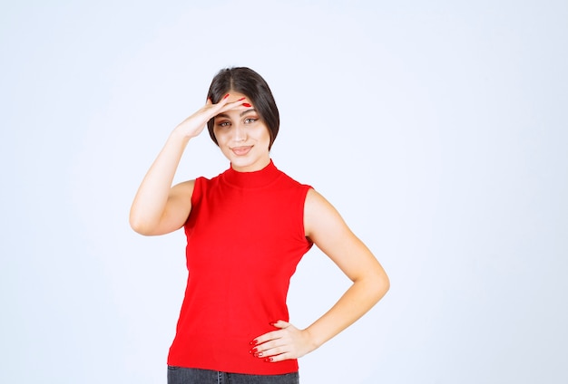 Girl in red shirt putting hand to her forehead and looking forward.