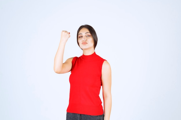 Девушка в красной рубашке показывает мышцы рук и кулаки.