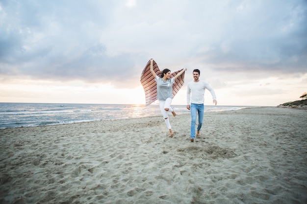 Girl running on the beach with a blanket and her boyfriend next to