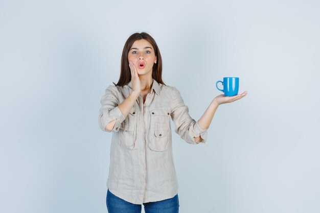 girl in shirt holding a cup of tea, putting hand on cheek and looking surprised.