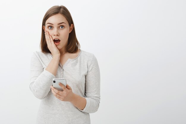 Girl shocked seeing embarrassing picture of friend in internet dropping jaw gasping from surprised holding palm on cheek staring amazed holding smartphone over gray wall