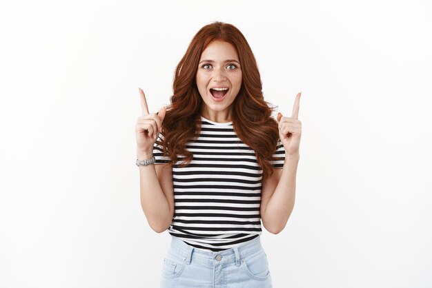 Girl show you must have product Enthusiastic charismatic redhead woman joyfully pointing top advertisement indicate up smiling amused with adoration and excitement white background