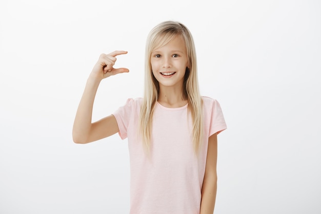 Девушка показывает, сколько усилий стоит, чтобы быть счастливым. Снимок в помещении: яркая дружелюбная белокурая девочка в розовой футболке, изображающая что-то маленькое или маленькое и широко улыбающаяся, возбужденная и радостная