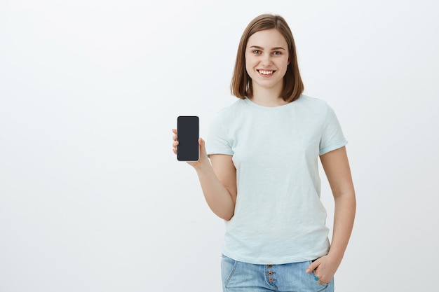 Girl showing new phone parents bought for new school term. Delighted and pleased charming young woman showing smartphone screen talking about cool app she found in online store over grey wall