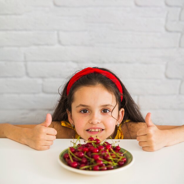 Girl showing thumb up sign in front of red cherries on plate over the desk