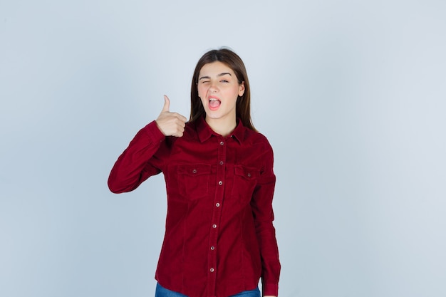 девушка показывает палец вверх, моргая в бордовой рубашке и выглядя довольной.