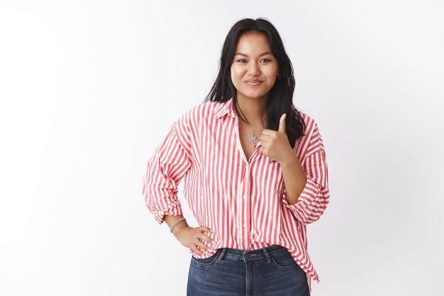 Girl showing thumbs-up delighted and pleased with awesome outcome of friend project smiling satisfied holding arm on waist assured, giving approval and positive opinion over white wall