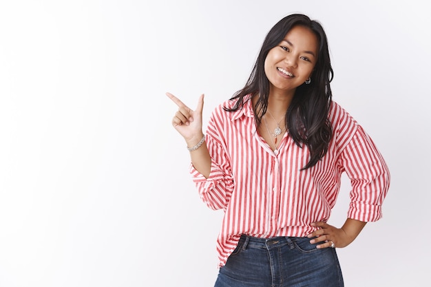 Girl shows best choice for you. Portrait of happy charming young 20s asian female holding hand on waist as pointing at upper left corner smiling and tilting head, suggesting awesome promo