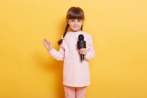 Girl singing with microphone and waving palm to camera, smiles, looking cute and charming, looks at camera, wearing casual clothing, kid with pigtails arranging concert for somebody.