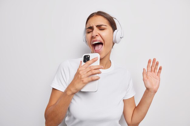 girl sings song listens music in headphones holds mobile phone dressed casually has cheerful expression on white