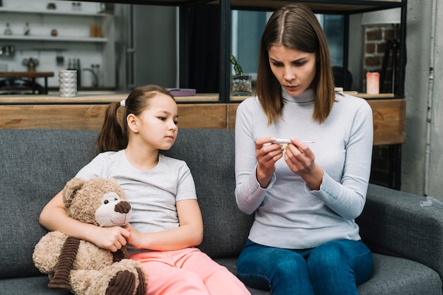 Girl sitting on gray sofa holding teddy bear looking at woman checking the temperature on thermometer