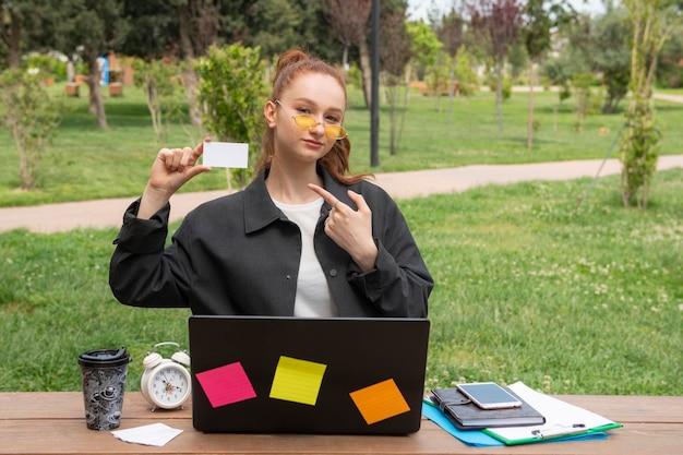 Girl sitting at laptop holding card pointing by finger smiling