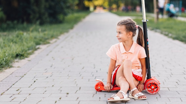 Girl sitting over push scooter on the straight walkway