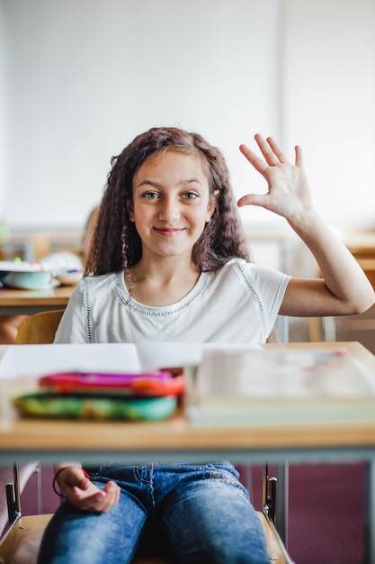 Girl sitting at school desk waving hand