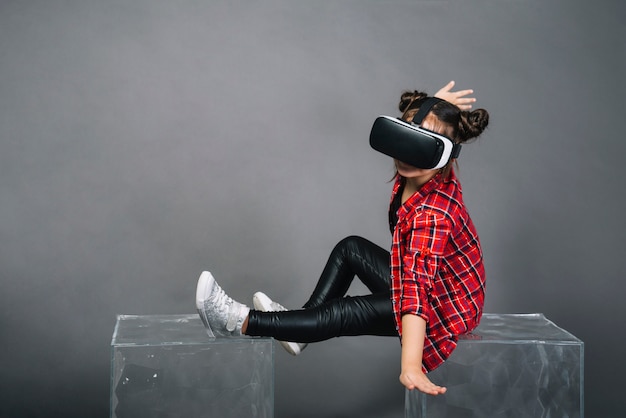 Девушка сидит на прозрачных блоках, носящих очки виртуальной реальности на сером фоне