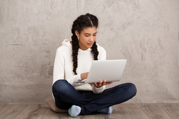 Girl smiling, holding laptop, sitting on floor over beige wall