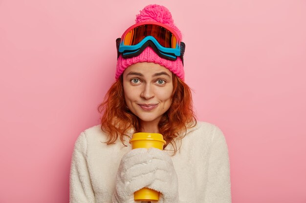 Girl snowboarder wears warm winter outfit, white mittens, holds takeaway coffee, isolated over pink background