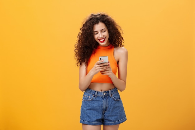 クロップドトップとデニムのショートパンツでオレンジ色の背景の上に幸せで明るい立っているデバイスの画面を見ながら笑っているスマートフォンを介してインターネットのテキストメッセージの友人のメッセージに時間を費やしている女の子。