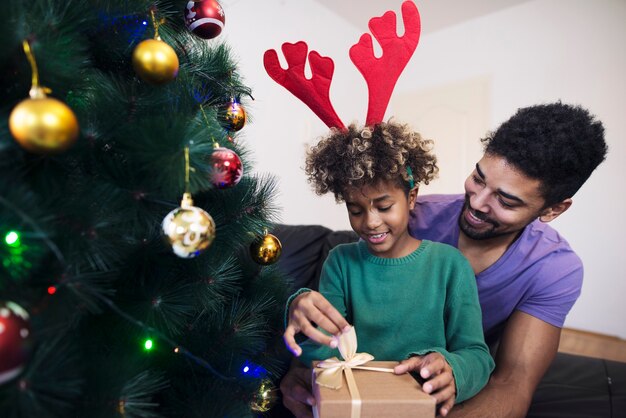 그녀의 아버지의 포옹에 크리스마스 트리 옆에 서서 선물 상자를 푸는 소녀