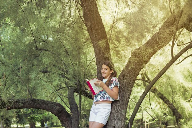 Girl standing by tree holding book smiling