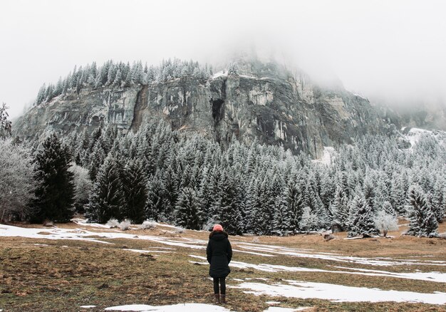 Girl standing in front of rocks and a forest covered in the snow under a cloudy sky