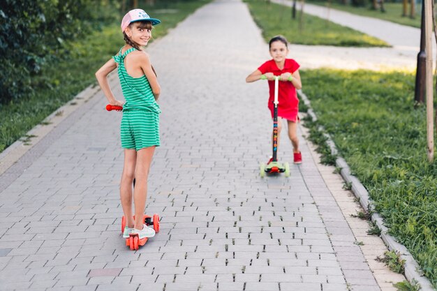 公園の歩道に乗っている彼女の友人とプッシュスクーターに立っている少女