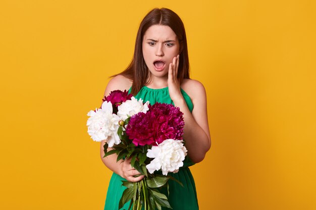 girl standing with bouquet of burgundy and white peonies in her hands, wearing green sundress, posing with open mouth, has astonished facial expression, isolated on yellow.