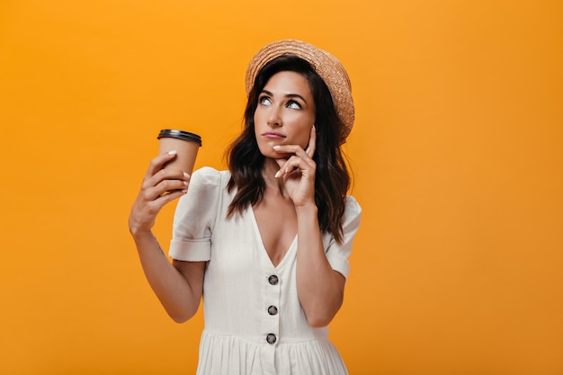 Girl in straw hat looks up thoughtfully and holds glass of coffee. Pensive woman in white summer clothes with coffee in her hands posing.