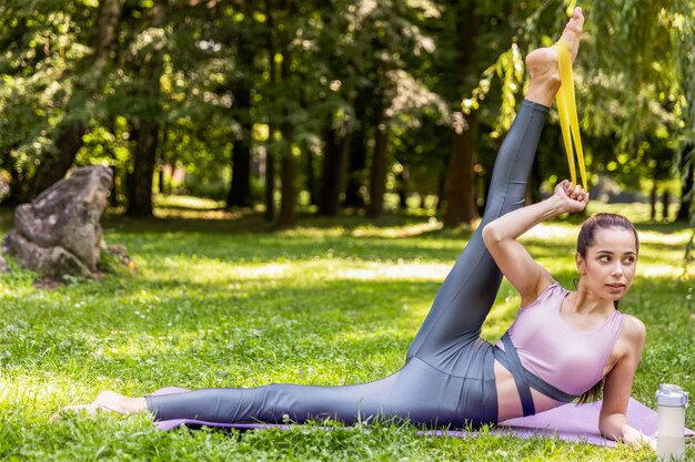 Girl stretching her foot with a yellow resistance band on the yoga mat in a park