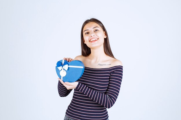 Девушка в полосатой рубашке держит синюю подарочную коробку в форме сердца и улыбается.