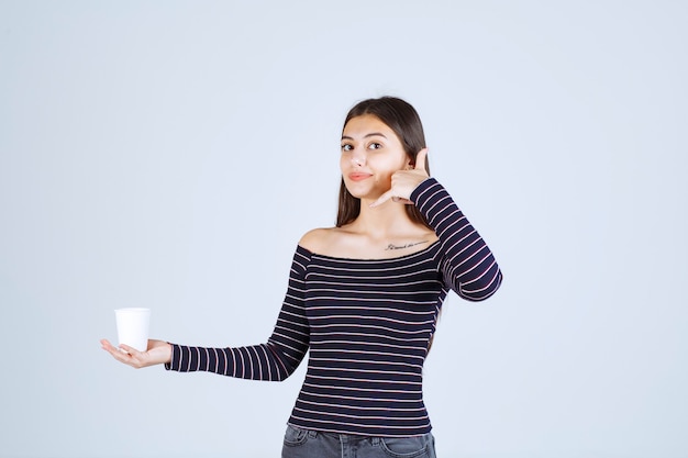 プラスチック製のコーヒーカップを持って電話を求めている縞模様のシャツの女の子。