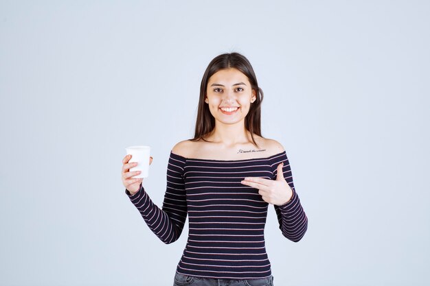 プラスチック製のコーヒーカップを持って、それを製品として紹介する縞模様のシャツの女の子。