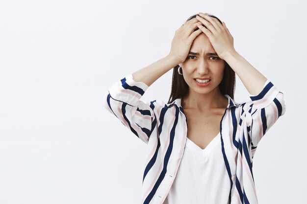 Girl stuck in terrible situation, thinking about way out. Portrait of troubled and stunned attractive female in striped blouse, holding palms on forehead, frowning and clenching teeth, being anxious