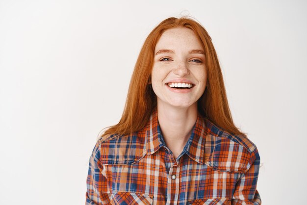 Девушка студентка с рыжими волосами и голубыми глазами счастливо улыбается в камеру Молодая рыжая женщина стоит весело на белом фоне