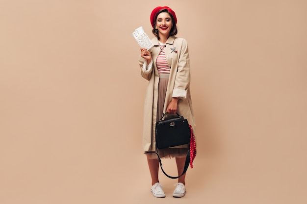 Girl in stylish outfit poses with handbag and tickets. Beautiful young woman in long beige skirt and coat smiles on isolated background.