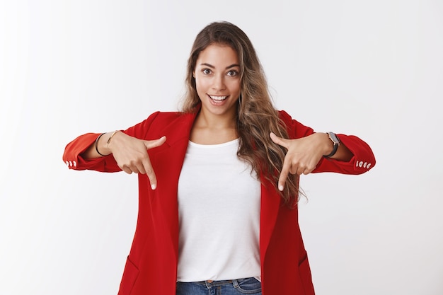 Girl suggest look downward great promotion raise company income. Excited good-looking assertive young female entrepreneur wearing red jacket pointing down smiling ambitious, white wall