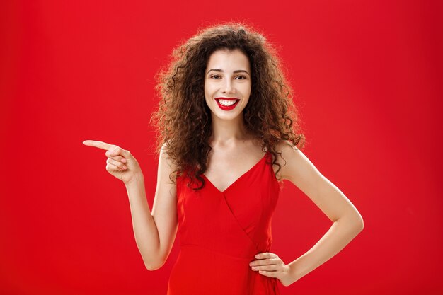 Girl suggesting ask her boyfriend. Charming elegant and stylish young female with curly hairstyle in red evening dress holding hand on hip and pointing left while showing friend where buy clothes.