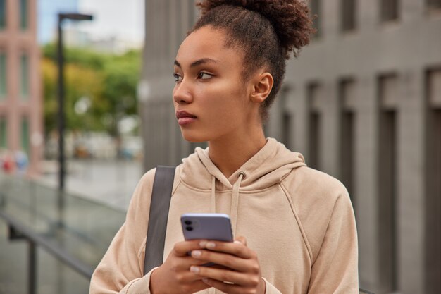 girl in sweatshirt holds digital modern mobile phone sends text messages carries karemat poses in urban setting rests after cardio training