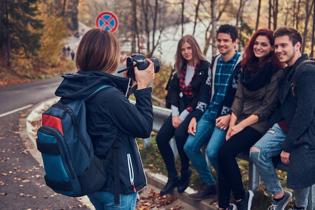 Девушка фотографирует своих друзей. Группа молодых друзей, сидящих на ограждении возле дороги. Путешествия, походы, приключения.