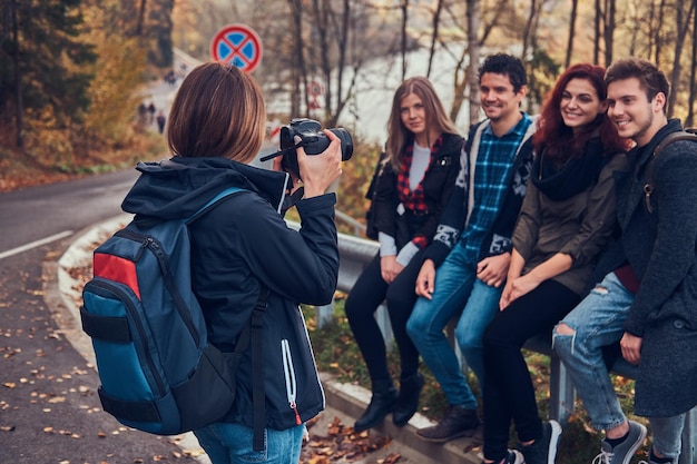 友達の写真を撮る女の子。道路の近くのガードレールに座っている若い友人のグループ。旅行、ハイキング、冒険のコンセプト。