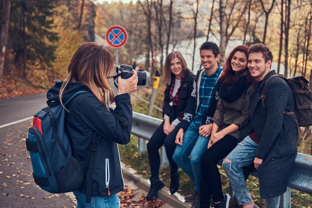 友達の写真を撮る女の子。道路の近くのガードレールに座っている若い友人のグループ。旅行、ハイキング、冒険のコンセプト。