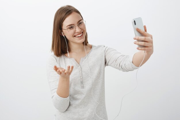 Девушка разговаривает с другом через видео-сообщение, жестикулируя рукой во время забавного разговора, указывая камерой смартфона на лицо в наушниках и улыбаясь экрану устройства над серой стеной