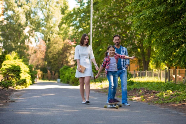 Девушка-подросток катается на скейтборде в городском парке. Родители смотрят на нее и улыбаются